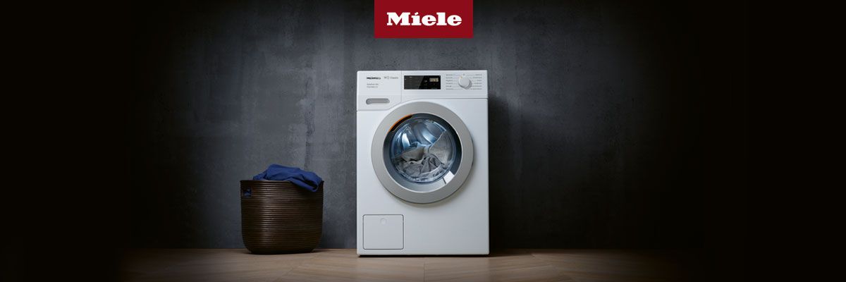 Weiße Waschmaschine vor dunkler Wand, daneben ein Korb mit blauer Kleidung. Oben rotes Miele-Logo, auf Holzfußboden. Weiße Waschmaschine vor dunkler Wand, daneben ein Korb mit blauer Kleidung. Oben rotes Miele-Logo, auf Holzfußboden.